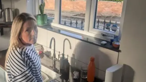 A woman in a black and white striped top stands in her kitchen next to her window, where you can see holes where a stone pavement previously was