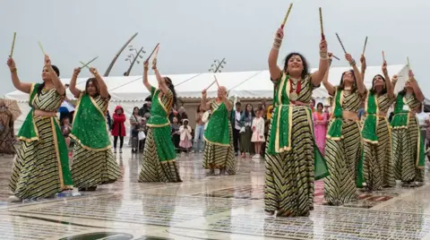 Caroline James Photography Indian dancers in two lines, in long, green dresses dancing on the comedy carpet in Blackpool, holding sticks above their heads.