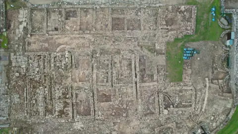Vindolanda Charitable Trust An aerial-view shot of the archaeological site, which shows the outlines of walls where buildings would've stood, closely packed together.