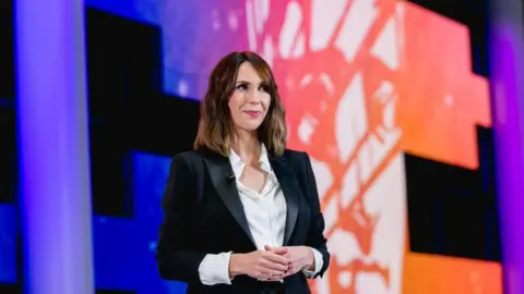 Getty Images Alex Jones is stood in front of a graphic in central square cardiff. she is wearing a suit