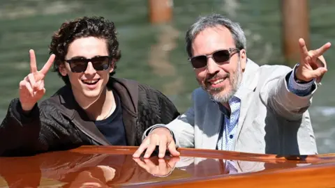 Timothee Chalamet and Denis Villeneuve together on a boat at the Venice Film Festival smiling and each flashing V peace sign with their hands