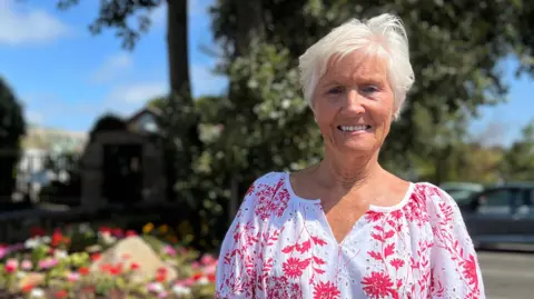Christine is standing beside a flower bed filled with colourful flowers near a white building and roadside trees. A large stone feature sits in the garden, with parked cars visible in the background.