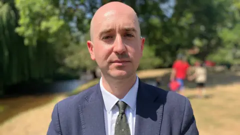 Owen Sennitt/BBC A man wearing a tie and suit looks towards the camera while standing on a village green next to a river.