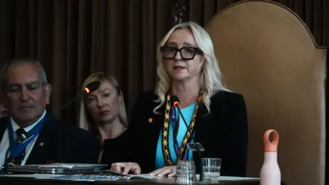 John Fairhall/BBC A woman with blonde hair, black-framed glasses, a turquoise dress and a dark-coloured suit blazer. She is sitting on a big brown chair, speaking into a microphone. There is a man and a woman on her right.