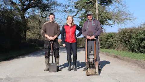 A group photo in Aldercar Lane. A middle-aged man is stood on the left with a shovel in front of him. A blonde-haired woman in a red coat is in the middle. Then an older man holding a wacker plate with a grey baseball cap is on the right.