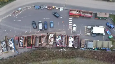 A drone shot of the Lower Compton recycling centre. A row of large metal skips can be seen from above, with parking spaces next to them and several cars parked up. There is a road next to it.
