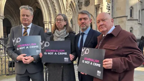 Vincent Kearney, wearing a dark grey suit, white shirt and a striped tie, stands outside the Royal Courts of Justice in London during an earlier hearing in the case in February. He is standing beside a woman and two men, one of whom is BBC Northern Ireland Director Adam Smyth. Three of them are holding black signs which read: "Journalism is not a crime."