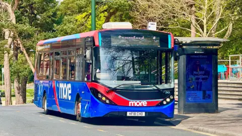 A blue and red single-decker bus with the More logo on the side. It's parked at a bus stop next to a park full of trees.