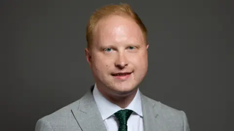 Man with short red hair and blue eyes smiles in grey suit, white shirt and green tie infront of grey portrait background