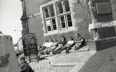 Alan Van Wijgerden The black and white image shows six men sitting in the sunshine outside Birmingham Gold and Silver Vaults. There is a sign that says such and another boy in the foreground