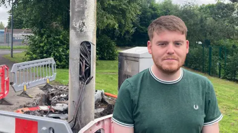 A man in a green shirt is stood in front of a metal lamp post. There is a hole cut out of the post with thick wiring protruding from the hole, it is scorched and badly damaged. 