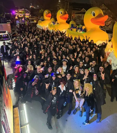 Stephen Rea A shot looking down on a crowd of people most dressed in black. Some have their arms up in the air. The crowd is in front of several large floats which look like yellow rubber ducks. 