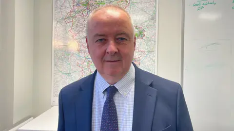 BBC A man with very short grey hair is wearing a dark blue suit jacket with a dark blue patterned tie, and white shirt with light blue squares on it. He stands in front of a whiteboard and a map of Staffordshire