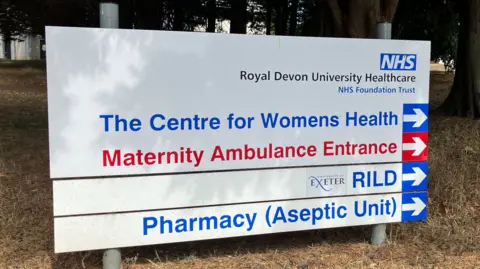 A hospital sign reads Royal Devon University Healthcare and directs people to The Centre for Womens Health, Maternity Ambulance Entrance as well as the RILD (Research Innovation Learning & Development) building and Pharmacy Aseptic Unit.