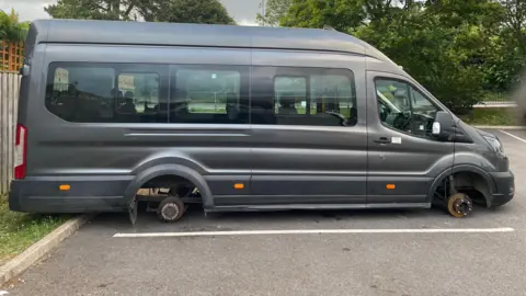 A dark grey Ford Transit van resting on the ground of a car park with its axles exposed having had its tyres stolen.