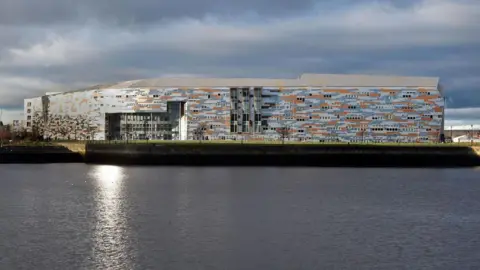 LDRS A general view of Middlesbrough College taken across the dock, with the water in the foreground. The large building is clad with grey, blue and orange material creating an irregular pattern.
