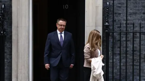 Getty Images A man in a suit is exiting the black door of 10 Downing Street. A woman is walking ahead of him, her face obscured by her long hair. 