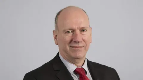 David Rees stood in front of a grey background, wearing a black suit, red tie and shirt.