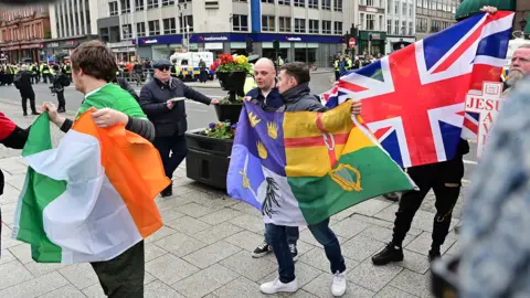 Pacemaker Six men hold up three flags in front of a large crowd of people. The flags, going from left to right, are an Irish tricolour, that off the 4 provinces of Ireland and a red, white and blue union jack