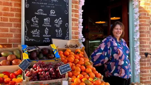 Karen Oakley Seorang wanita berdiri di pintu sebuah toko, dengan kotak buah dan papan daftar pilihan makanan.