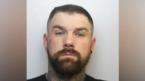 Avon and Somerset Police A mugshot of a man in his 30s with dark hair and a beard. He has tattoos all over his neck as well as one on one of his cheeks.