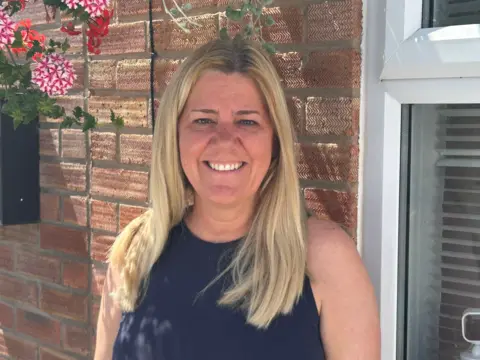 BBC / Gavin Kermack A woman with long blonde hair wearing a blue sleeveless top, standing in front of a brick wall with a hanging basket attached to it.