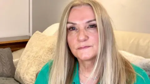 A woman with blonde hair, wearing a green top, sat on a sofa and looking into the camera