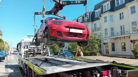 A red car is being lifted onto a flatbed truck. The is a man in an orange florescent jacket standing next to the white truck. It is a sunny day with a blue sky and the truck is parked on a residential street where you can see a four-storey building. The road has double yellow lines. 