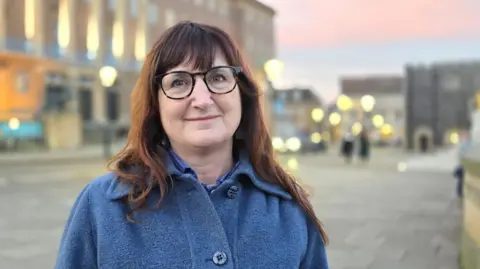 Paul Moseley/BBC Lucy Galvin is looking at the camera. She has long brown hair and glasses and is wearing a blue fabric coat. She is standing in front of Norwich City Hall, which out of focus in the background.