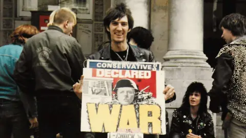 Simon Masterson A man with long black hair wearing a leather jacket holding up a protest placard that says WAR on it