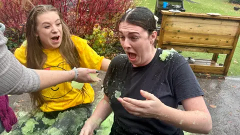 Two women covered in green gunge. Heidi Simmons, on the left, has her mouth open but is smiling a little. She is sat next to Ella Samson, who has her mouth open and a frown on her face in shock. 