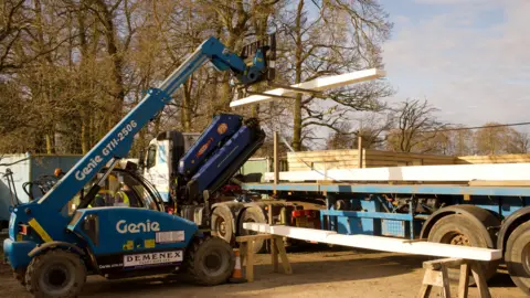 A blue forklift truck lifts wood on to a flat bed lorry in a yard.