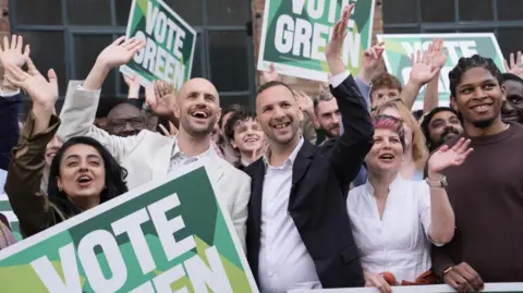 Green Party of England and Wales leader Zack Polanski stands in the middle of local election candidates, some holding placards with the large slogan "Vote Green". He wears a black jacket and a beige, open-necked shirt. 