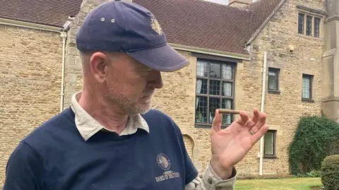 Man dressed in a blue top and blue cap looking at a wartime bullet he is holding in his left hand.