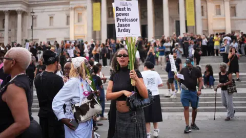 A crowd of people gather in Trafalgar Square as part of a vigil for Jason Arday. Some of them are holding placards.