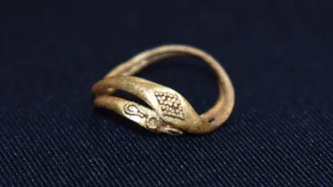 Colchester Archaeological Trust A pair of conjoined Roman gold rings, sitting on a piece of dark material. One ring is decorated with 25 tiny gold pellets arranged in a diamond shaped pattern. The other has a flat, elongated bezel decorated with fine strands of gold wire forming a central circle with drop-shaped motifs on either side, possibly representing a flower. Together, they are delicate, skilfully made objects, dating to between the 1st and 3rd centuries AD.