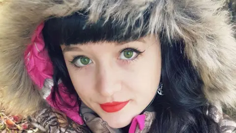 A close-up selfie of a woman with black hair looking straight at the camera. She has green eyes and is wearing red lipstick. The fur lining of a hood can be seen over her head.