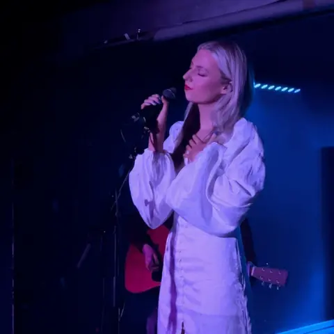 Riley music Riley - a young woman standing on stage, holding a microphone. She is wearing a long white dress with puffed bell sleeves and red lipstick. Her eyes are closed as she sings and she is illuminated by a blue spotlight in a dark venue - a musician with an acoustic guitar is visible behind her