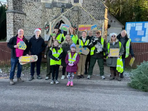 Nancy Bassant A group of adults and children wearing high-vis jackets stand outside a church, some holding homemade placcards