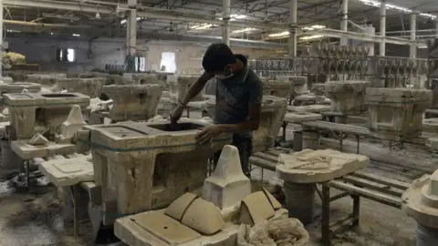 A man works on a toilet seat at a ceramic factory in Gujarat's Morbi in November 2016. He is in a large factory hall, surrounded by a large number of finished and unfinished sanitaryware.