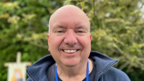 Andy Leary has an excited smile, he has blue eyes and is wearing a dark blue coat. He's standing in front of a green bush which is blurred.