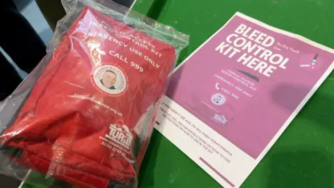 A red fabric bag, containing medical equipment for use in the event of a stabbing. It has white writing on saying it is for emergency use only and to call 999. It has an image of Daniel Baird in the centre on a white background. A piece of paper next to it has details about the kit. 