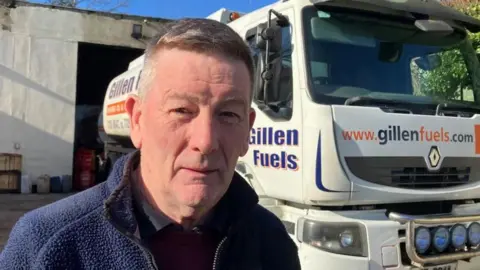 Anthony Gillen is looking at the camera. He is wearing a navy fleece zip up. He is standing in front of an oil delivery truck with the family name one it.