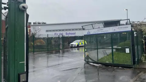 A photo taken from the school gates shows a police car parked in front of a school building. Purple letters on the white building read "Fir Vale School", although the E has fallen off.