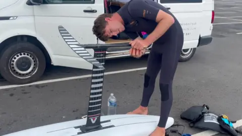 Emma Bridge A man in a wetsuit leans over an upside down hydrofoil board carrying out maintenance on it