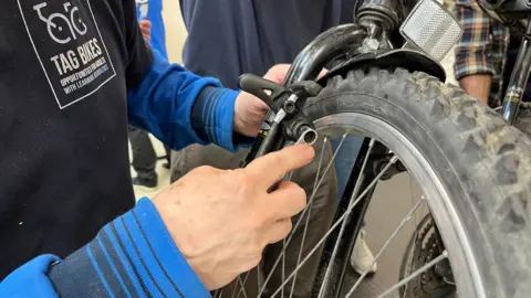 Tom Jackson/BBC A person is holding a wheel of a bike and is using a tool to tighten a screw. The person is on the left and is wearing a blue jumper with a navy T-shirt over the top which says TAG BIKES.
