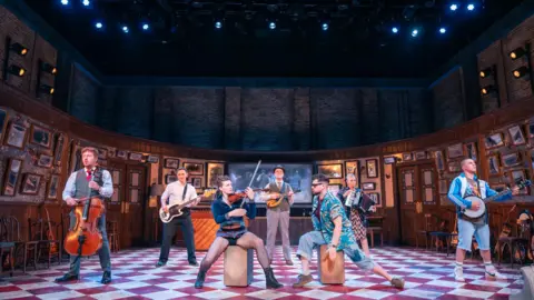 Tommy Ga-Ken Wan A group of performers play instruments and sing on stage in a pub setting during a production of Once.