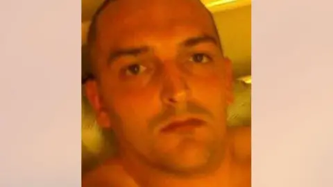 Paul Foster, who has a shaved head and is shirtless, looks into the camera with a serious expression.