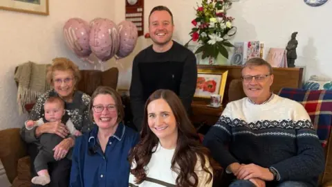 Davinia Ramos/BBC Three women and two men sit in a living room smiling at the camera. An old woman holds a young baby on her lap. 