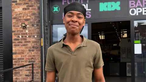 HarvStar - wearing a black cap and a green shirt - smiles as he stands outside the front entrance of a building.